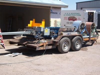 Metal roofing machine on a trailer outside a building; a roll of metal sits on one side.