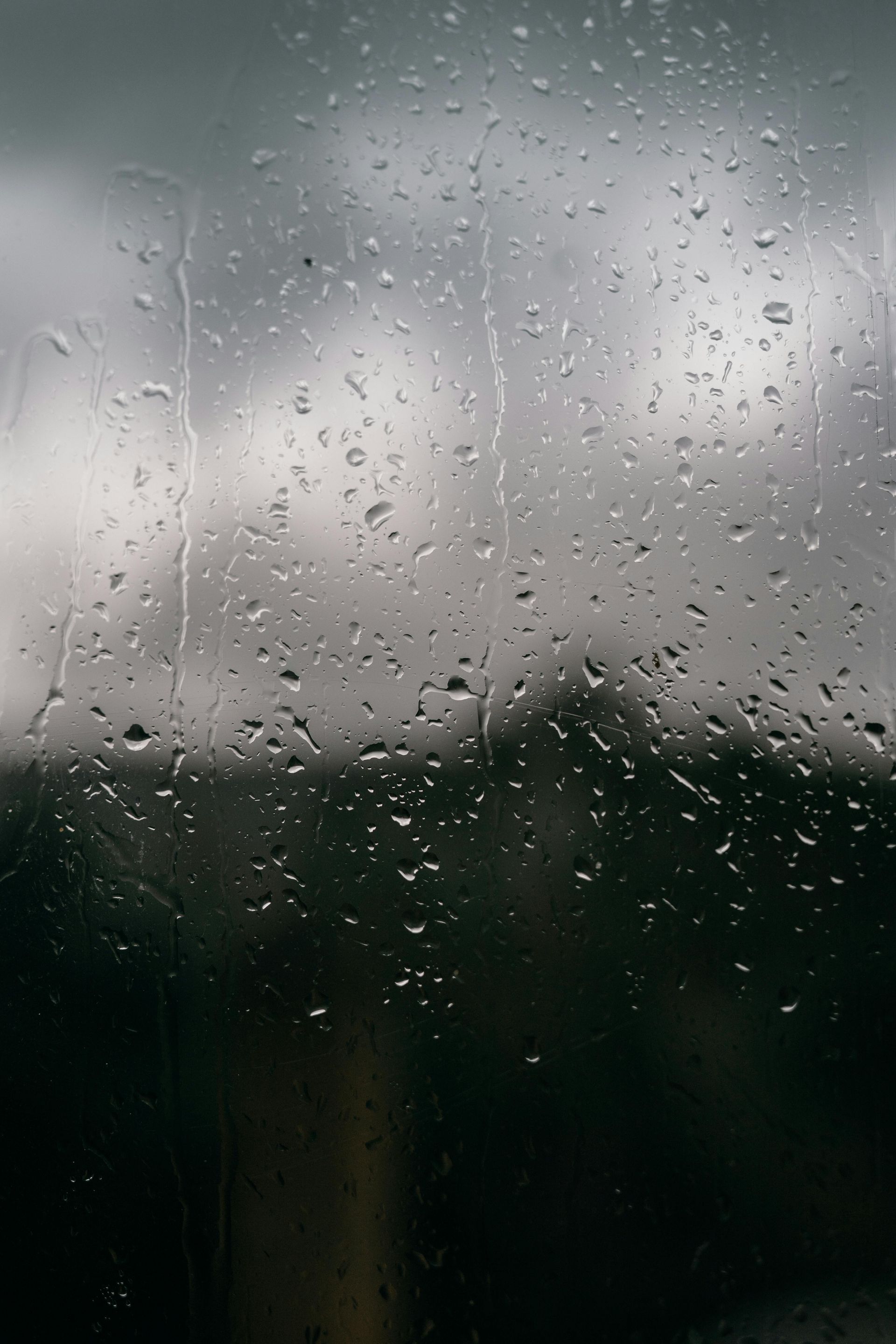 Raindrops on a window, blurred dark background with hints of trees and overcast sky.