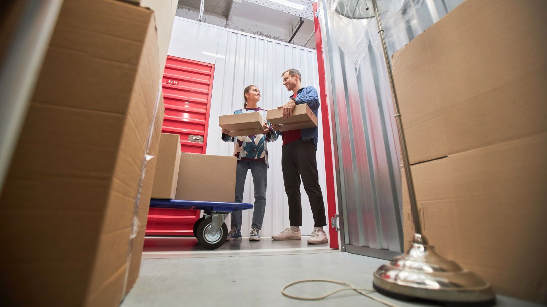 Deux personnes transportant des boîtes dans une unité de stockage avec des boîtes, un chariot et une lampe.