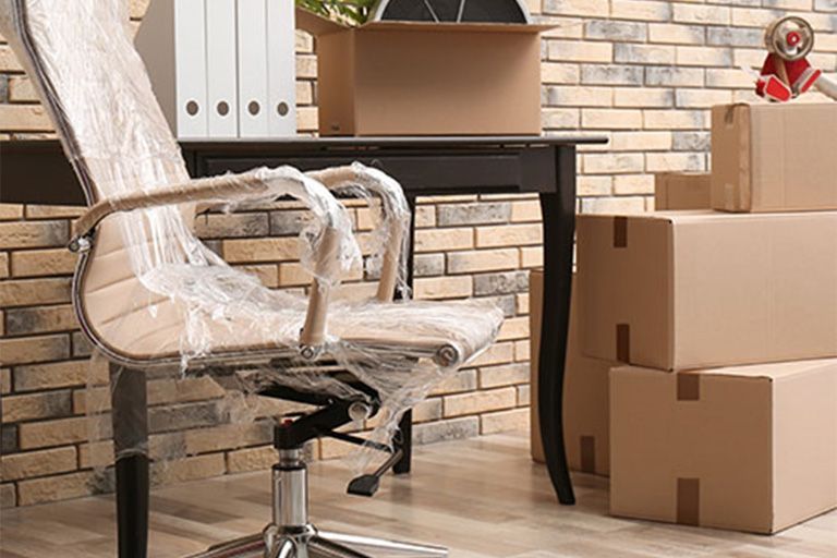 Chaise de bureau enveloppée dans du plastique, bureau et cartons contre un mur de briques, indiquant un déménagement de bureau.