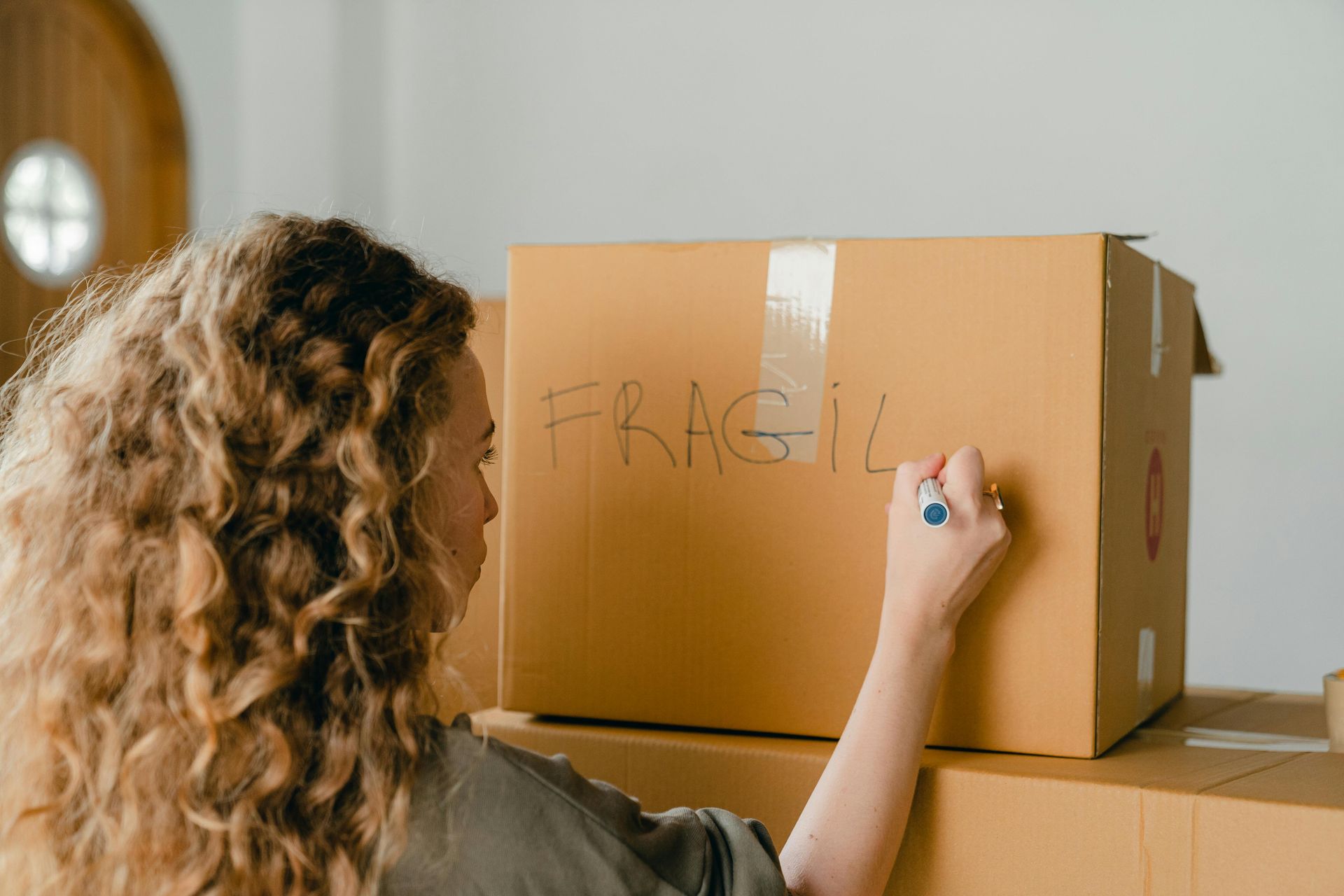 Dans une pièce lumineuse et peu meublée, une personne aux cheveux bouclés écrit « FRAGILE » sur un carton de déménagement brun.