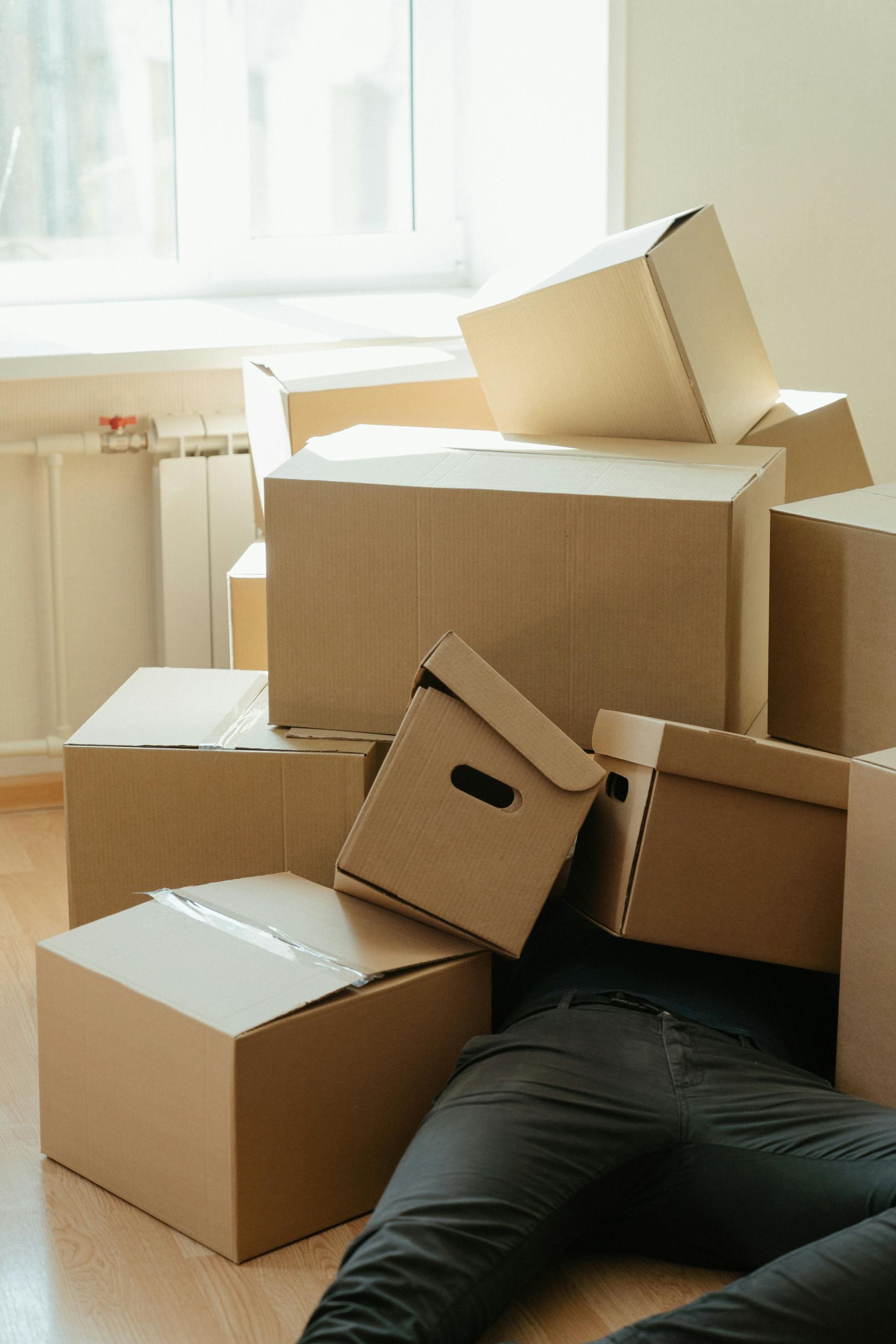 Dans une pièce lumineuse, une personne vêtue de noir est allongée sur le sol, dissimulée par une grande pile de cartons de déménagement bruns.