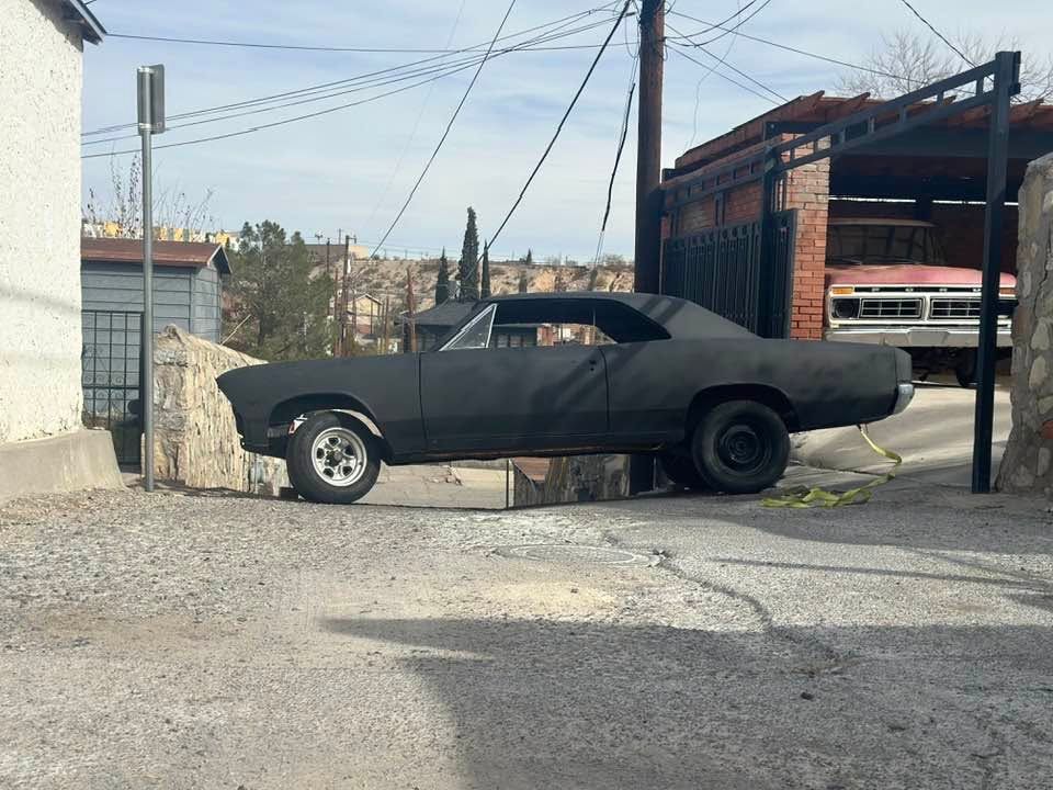 Black classic car parked in a gravel yard beside a red brick garage and utility poles | Pro Veterans Automotive