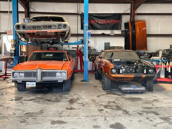 Three classic cars in a garage, including a lifted orange car and two cars on lifts.