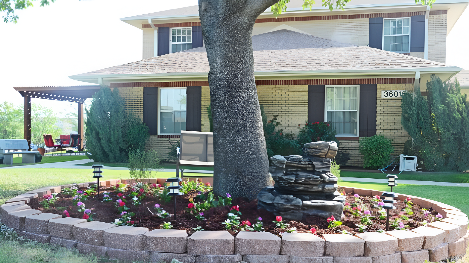 A circular stone garden bed surrounds a large tree in front of a tan house, featuring fresh mulch, flowers, and a fountain.