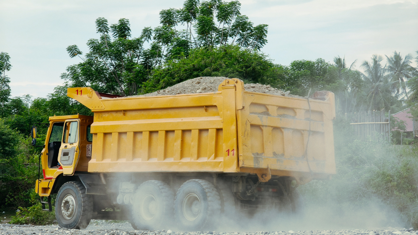A yellow dump truck driving on a gravel road, kicking up dust in a green, wooded area.