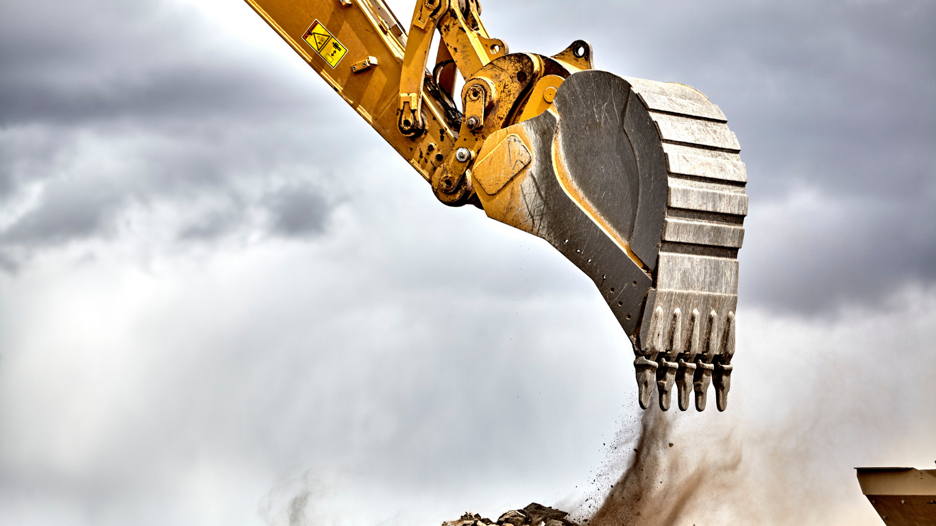 A yellow excavator bucket dumps dirt against a backdrop of a cloudy, overcast sky.