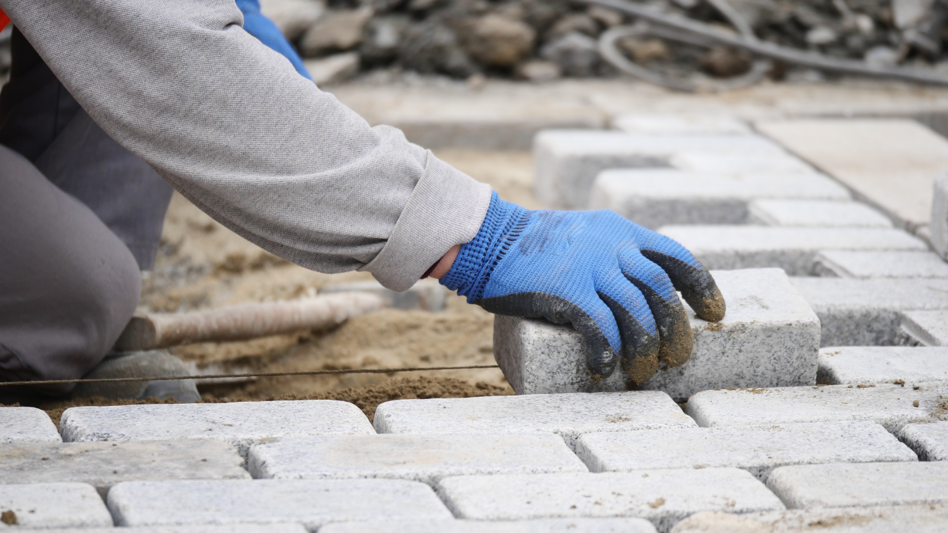 A person wearing blue gloves places a grey paving stone into a new walkway.