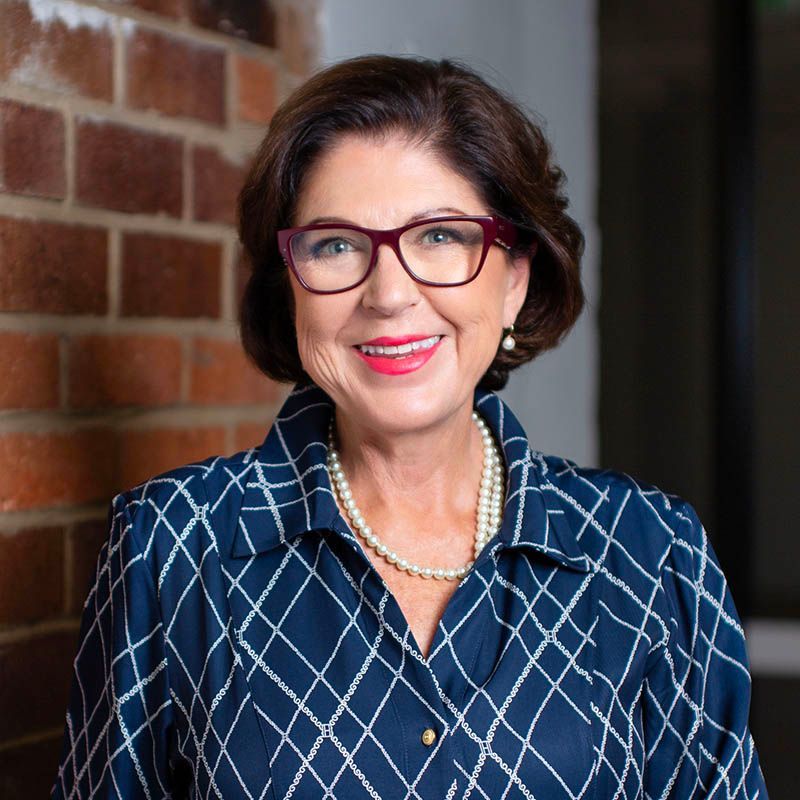 Smiling woman in red glasses and blue patterned jacket against a brick wall