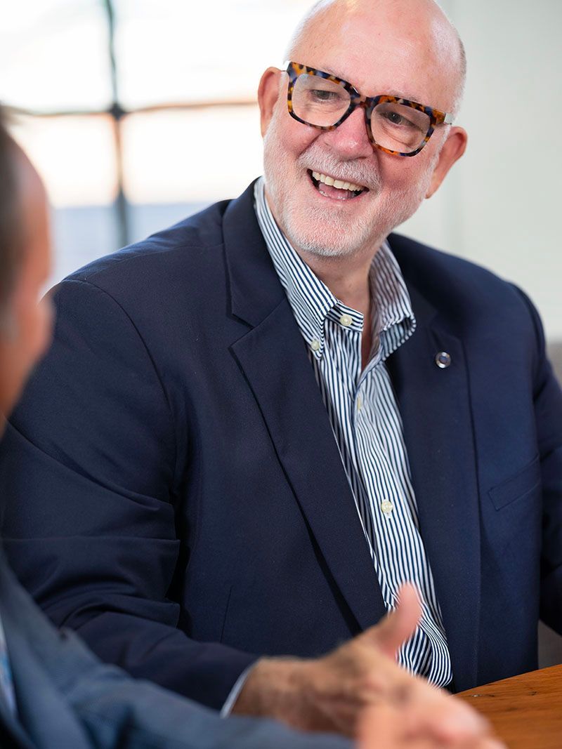 Smiling man in glasses and navy blazer giving a thumbs-up at a table.