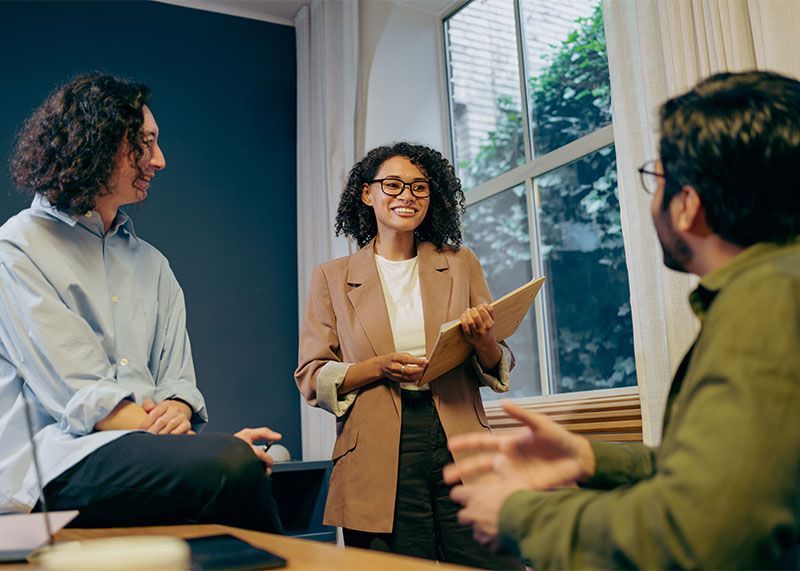 Three people in a meeting room, one woman presenting notes by a window.
