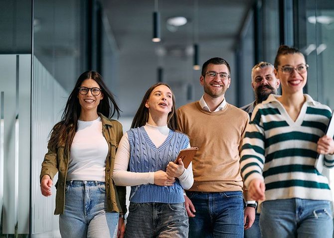 Five coworkers walking down a glass office hallway, smiling and chatting.