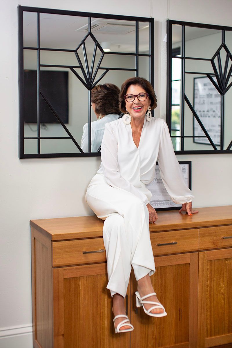 Woman in white outfit sitting on a wooden cabinet in a bright room with framed mirrors.