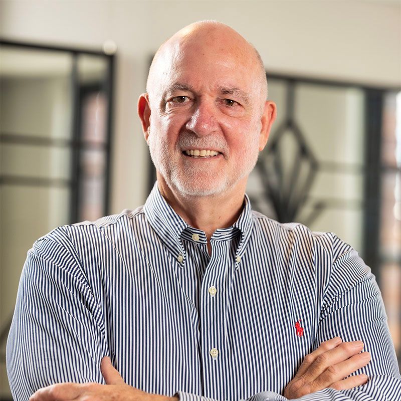 Smiling bald man in a striped shirt with arms crossed, standing indoors in a blurred office setting