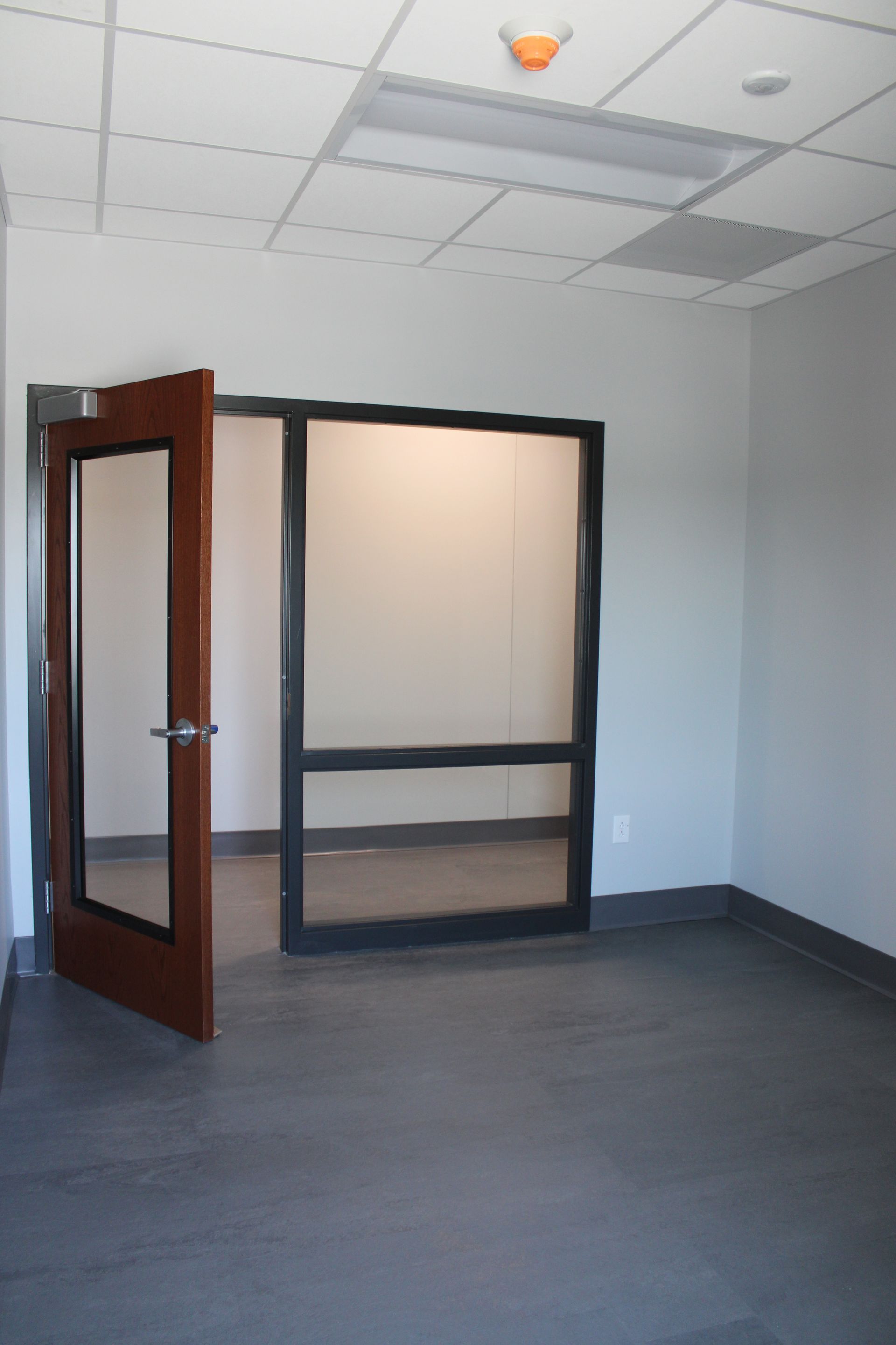 Empty office room with an open wooden door, gray carpet, and a glass partition wall.