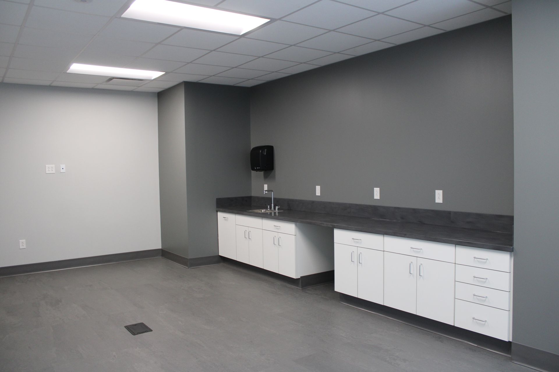 Empty break room with gray walls, white cabinets, and a long counter under fluorescent lights