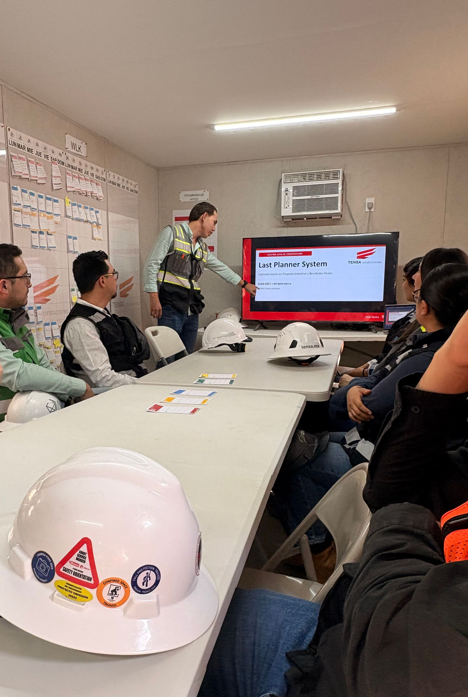 Team meeting in a conference room, with people watching a presentation on a monitor and a white hard hat on the table.