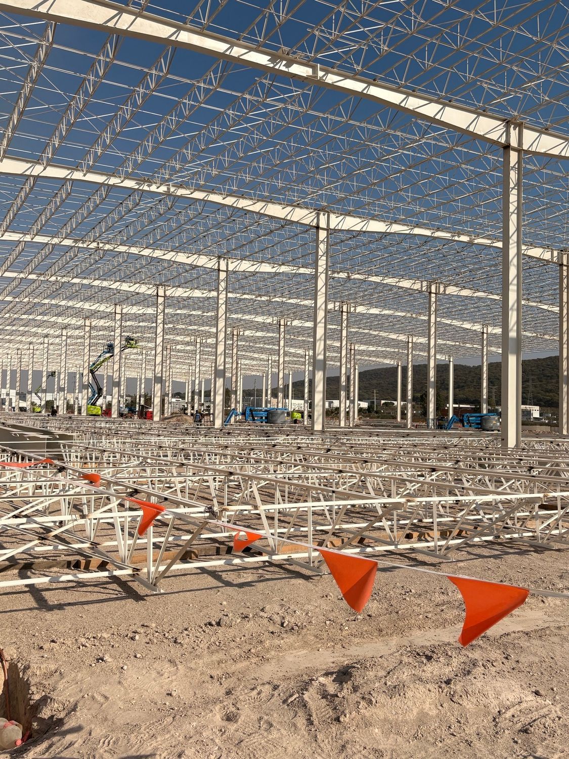 Construction site with a large steel frame, orange flags, and sandy ground under a blue sky
