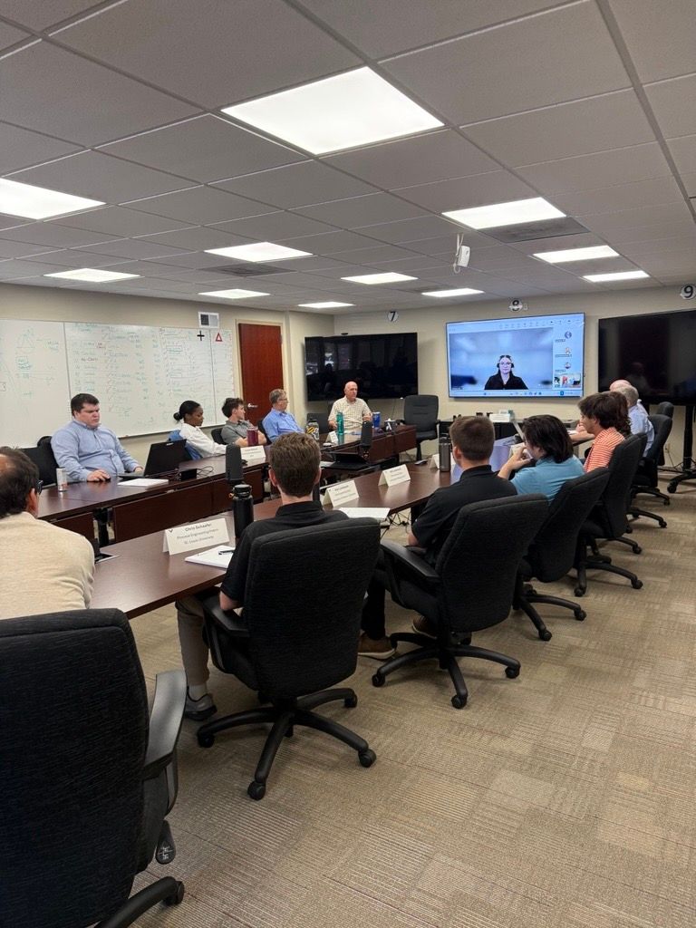 Conference room meeting with people seated around a table, viewing a presentation on a large screen.