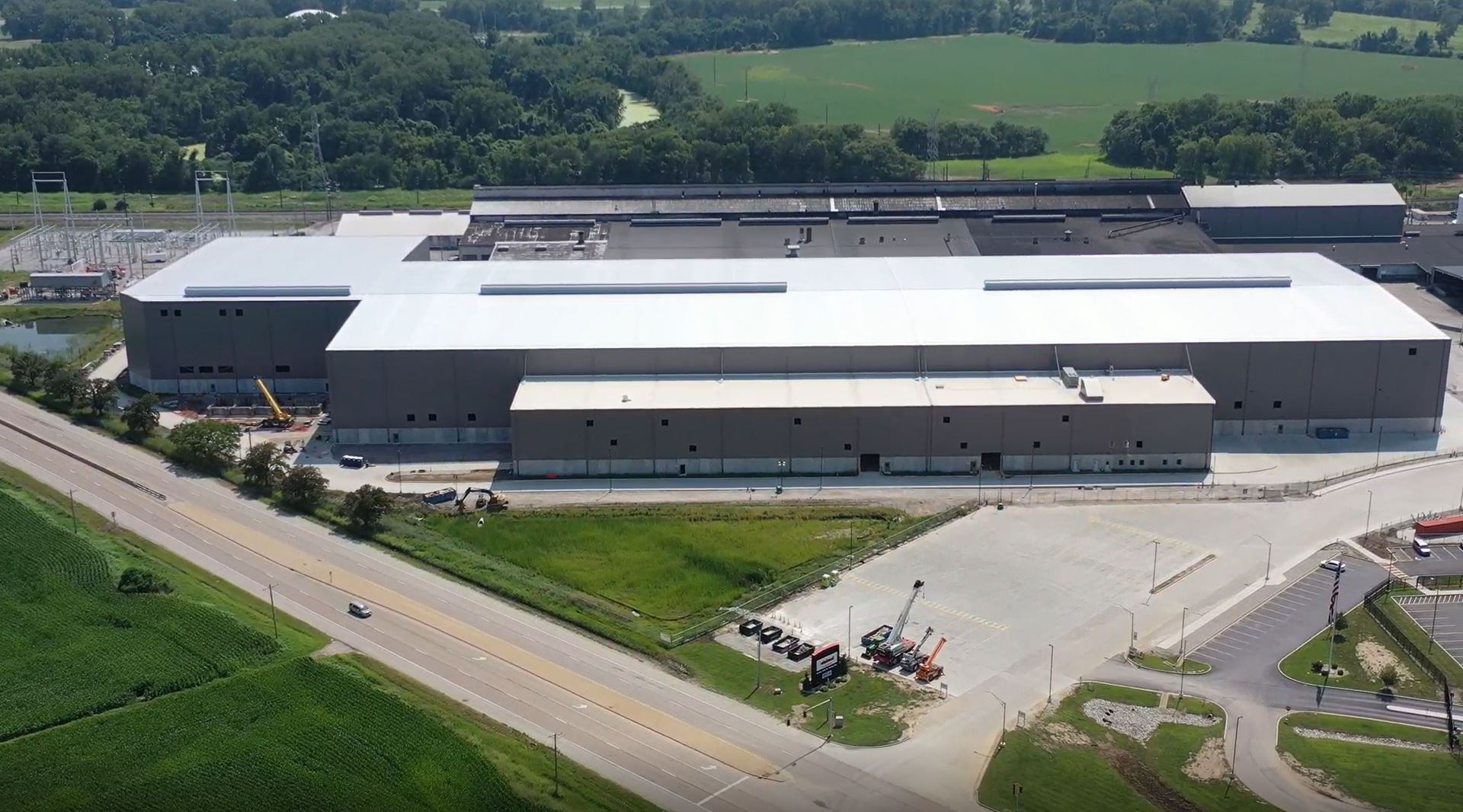 Aerial view of a large gray industrial warehouse beside a road, with parking lot and green fields nearby