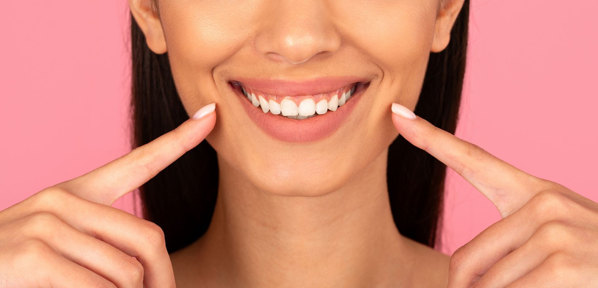 Woman smiling and pointing at her bright white teeth against a pink background