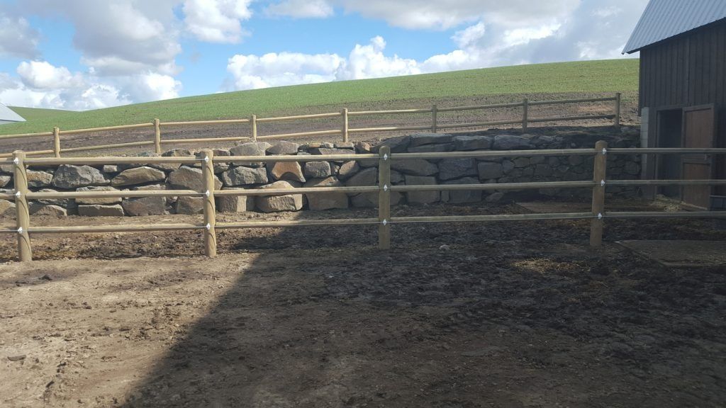 A wooden fence surrounds a dirt field next to a stone wall.