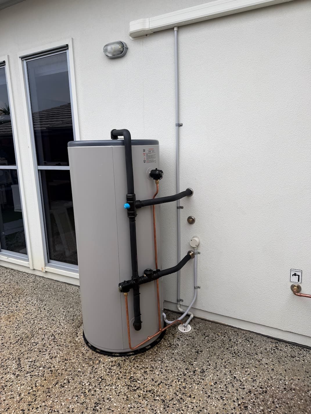 Outdoor water heater tank beside a white house wall near a glass door and gravel patio.