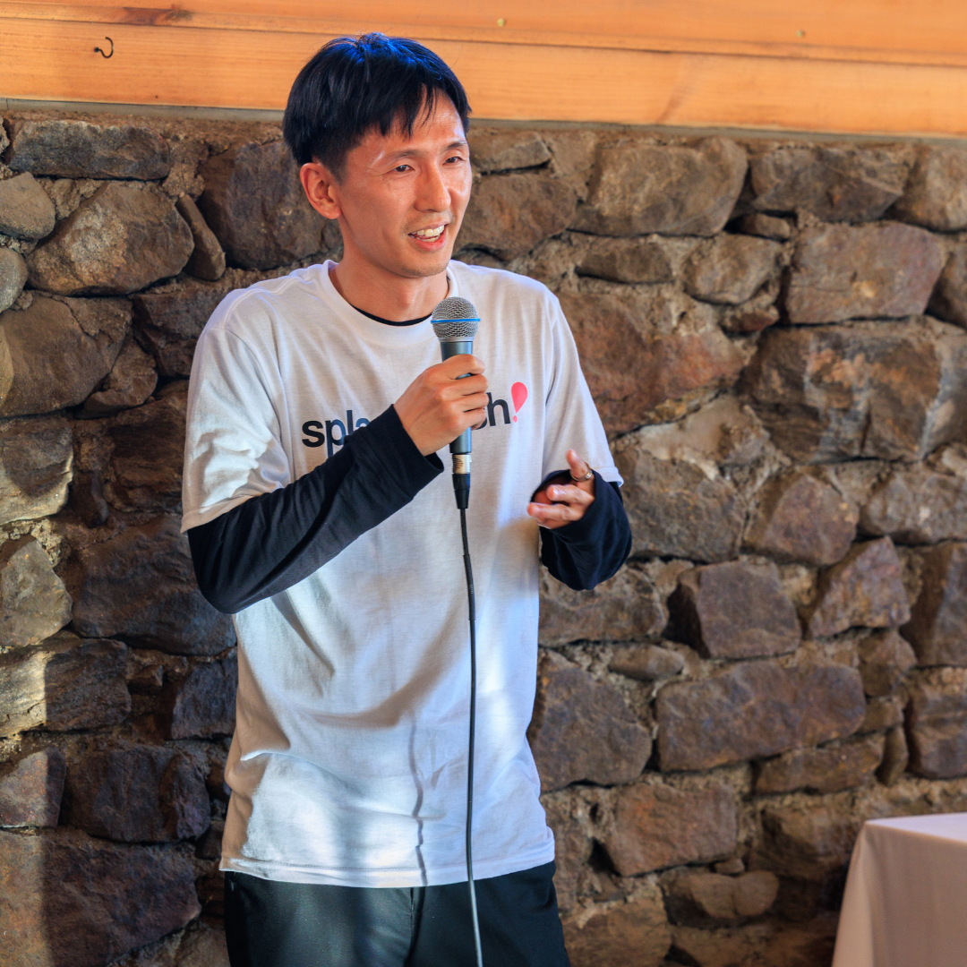 Man speaking into a microphone, smiling, standing in front of a stone wall, wearing a t-shirt.