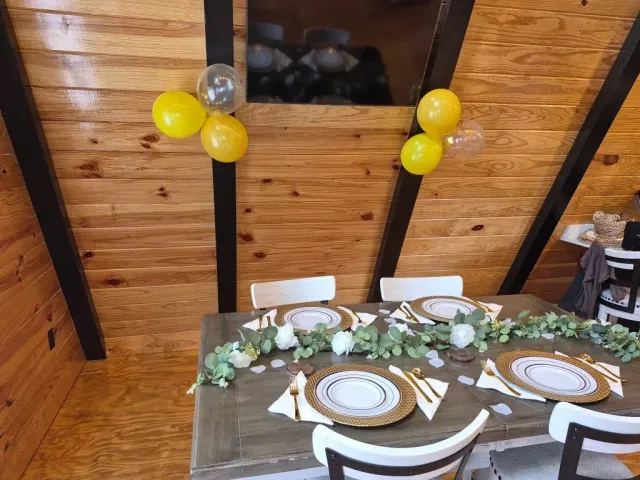 Rustic wedding table with white chairs, greenery centerpiece, and yellow balloons against a wood-paneled wall