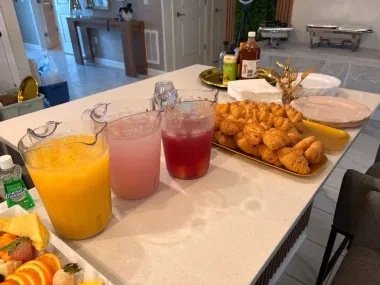 Pitchers of orange, pink, and red drinks beside a platter of fried snacks on a dining table.
