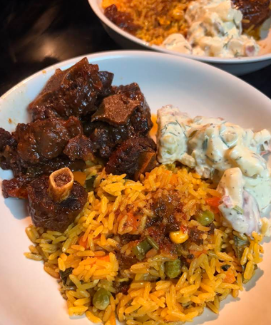 Plate of rice, dark stew meat, and creamy salad on a white dish, with another plate blurred in back