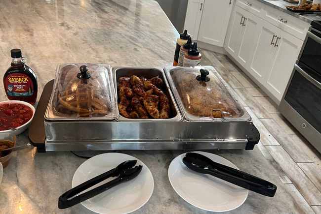 Buffet table with trays of food, sauces, plates, and tongs in a kitchen serving area
