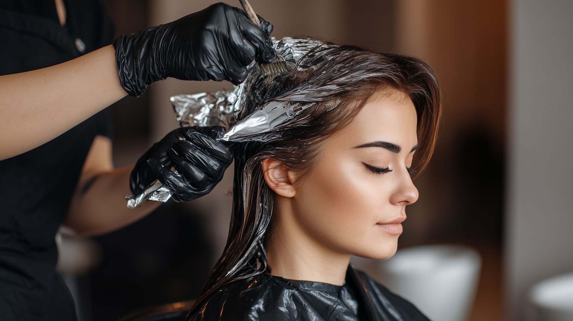 Hair colorist applying foil to a client's dark hair in a salon, client seated with eyes closed.