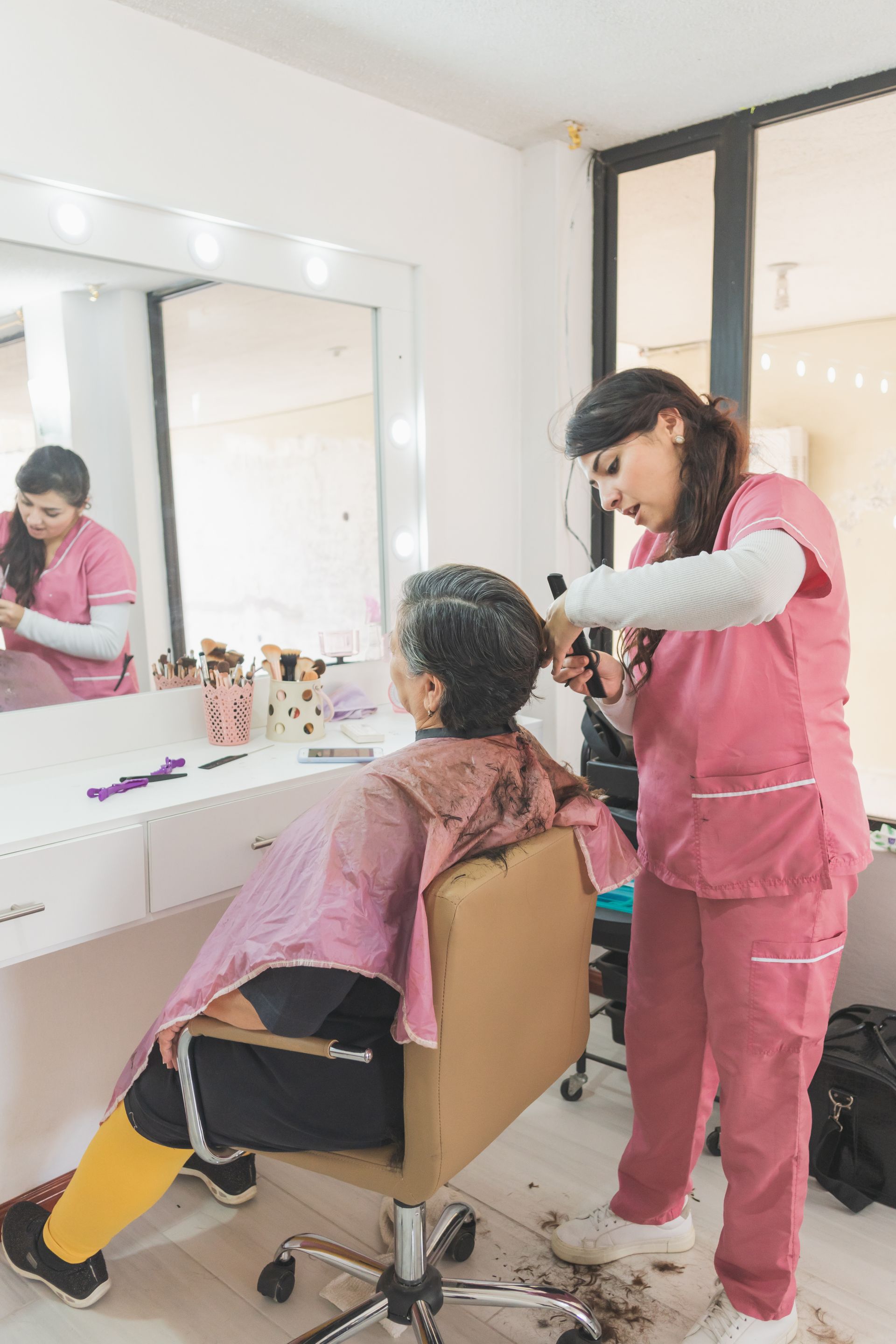 Hairdresser cutting a client’s hair in a salon, with mirror, styling chair, and bright pink smocks