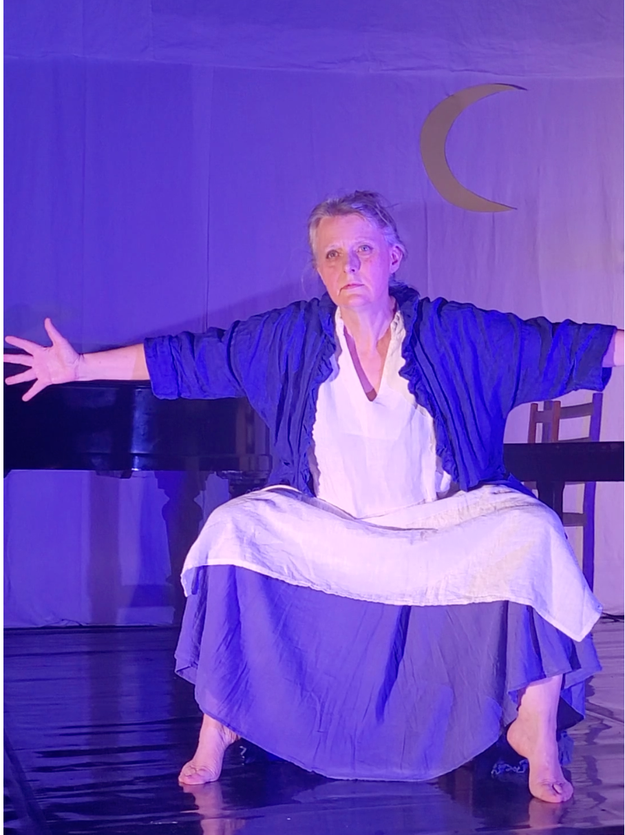 Femme en costume bleu et blanc, bras ouverts, accroupie sur scène sous un croissant de lune.