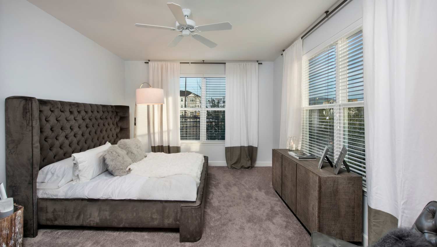 Bedroom with a dark tufted bed, white bedding, and large windows with white curtains at Olympus Preserve at Town Center.