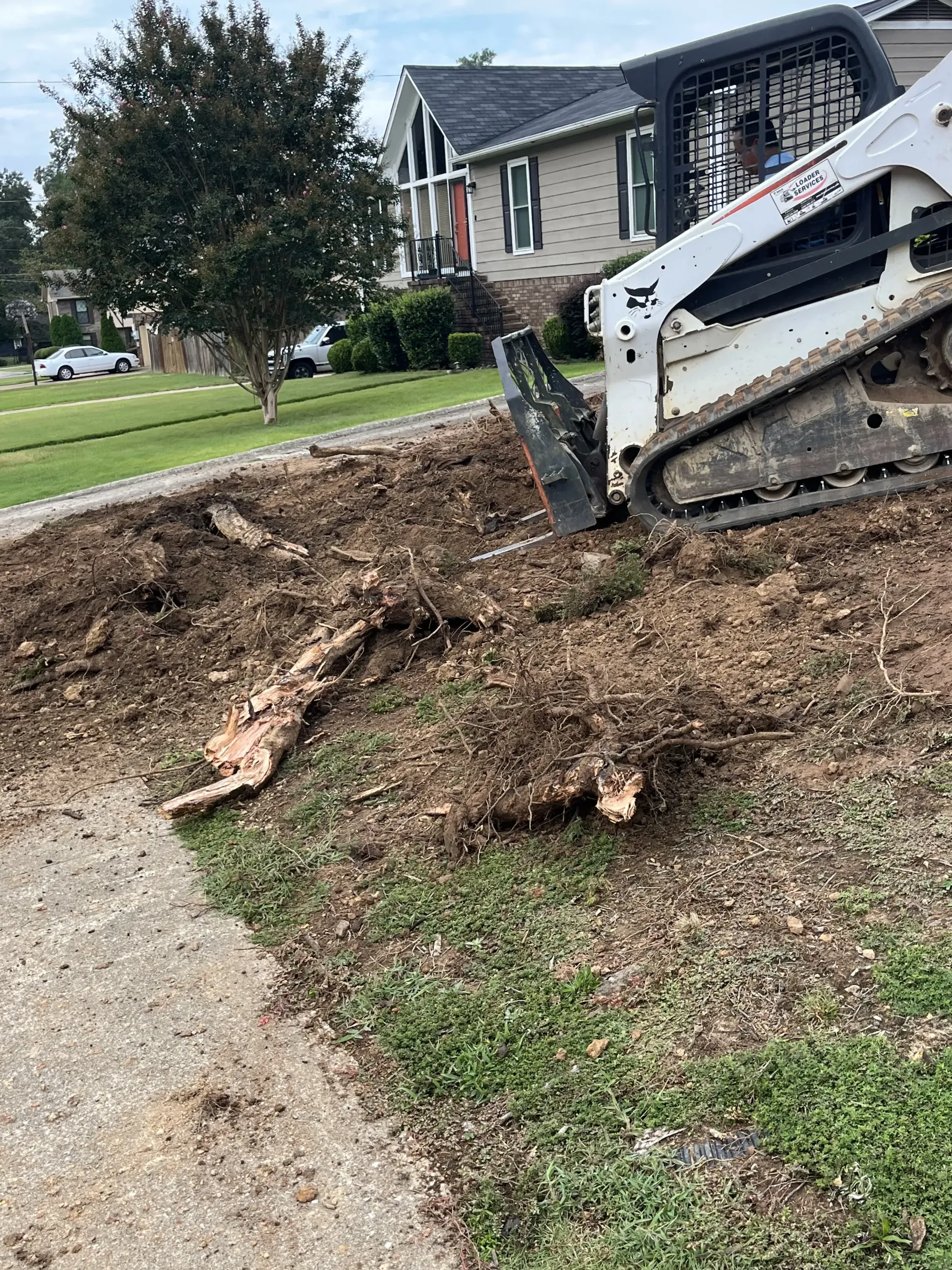 Bobcat digging a trench beside a suburban house, with dirt piled along a gravel edge.