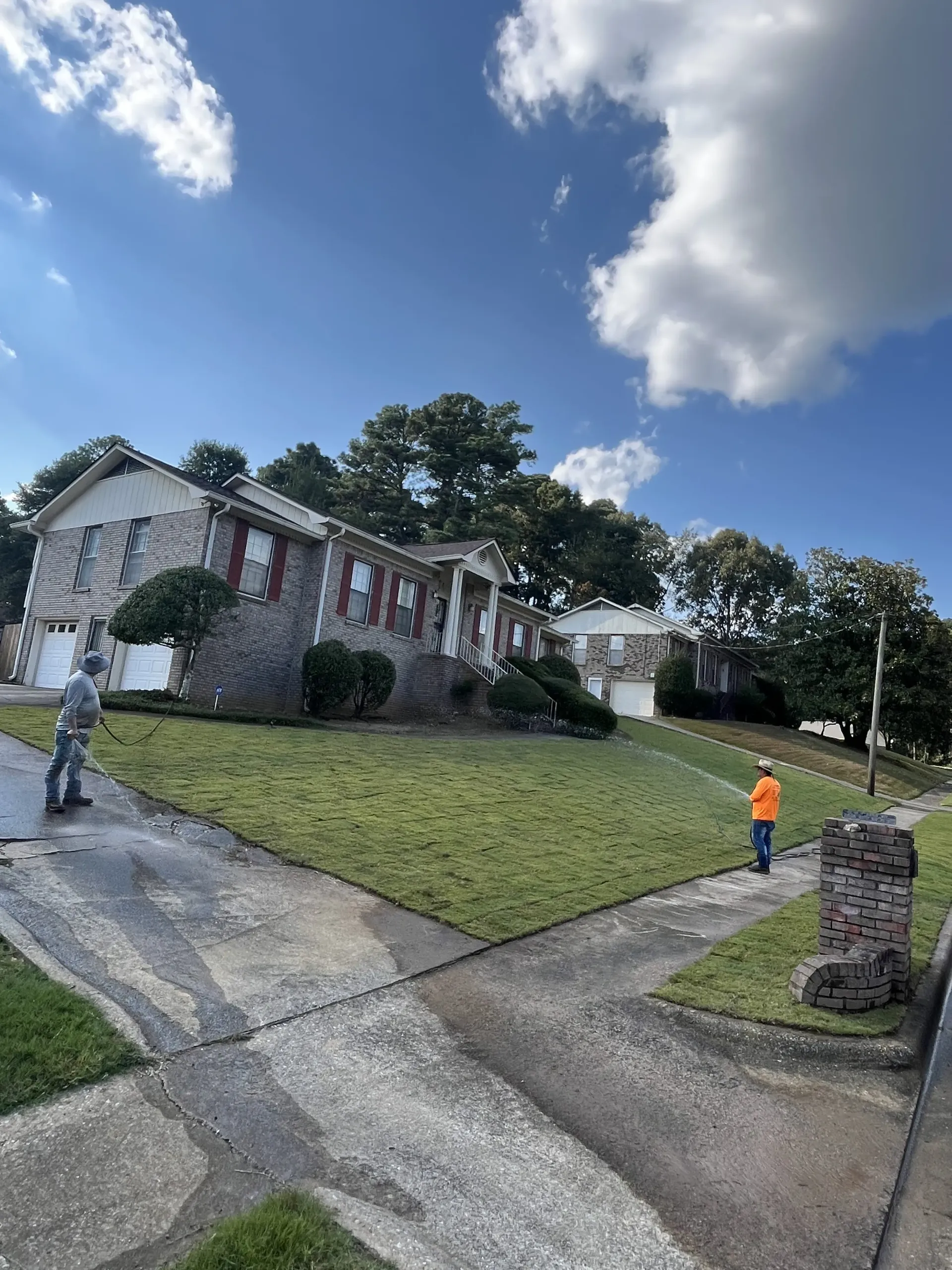 Suburban house with large lawn under a blue sky, with two people standing near the driveway.