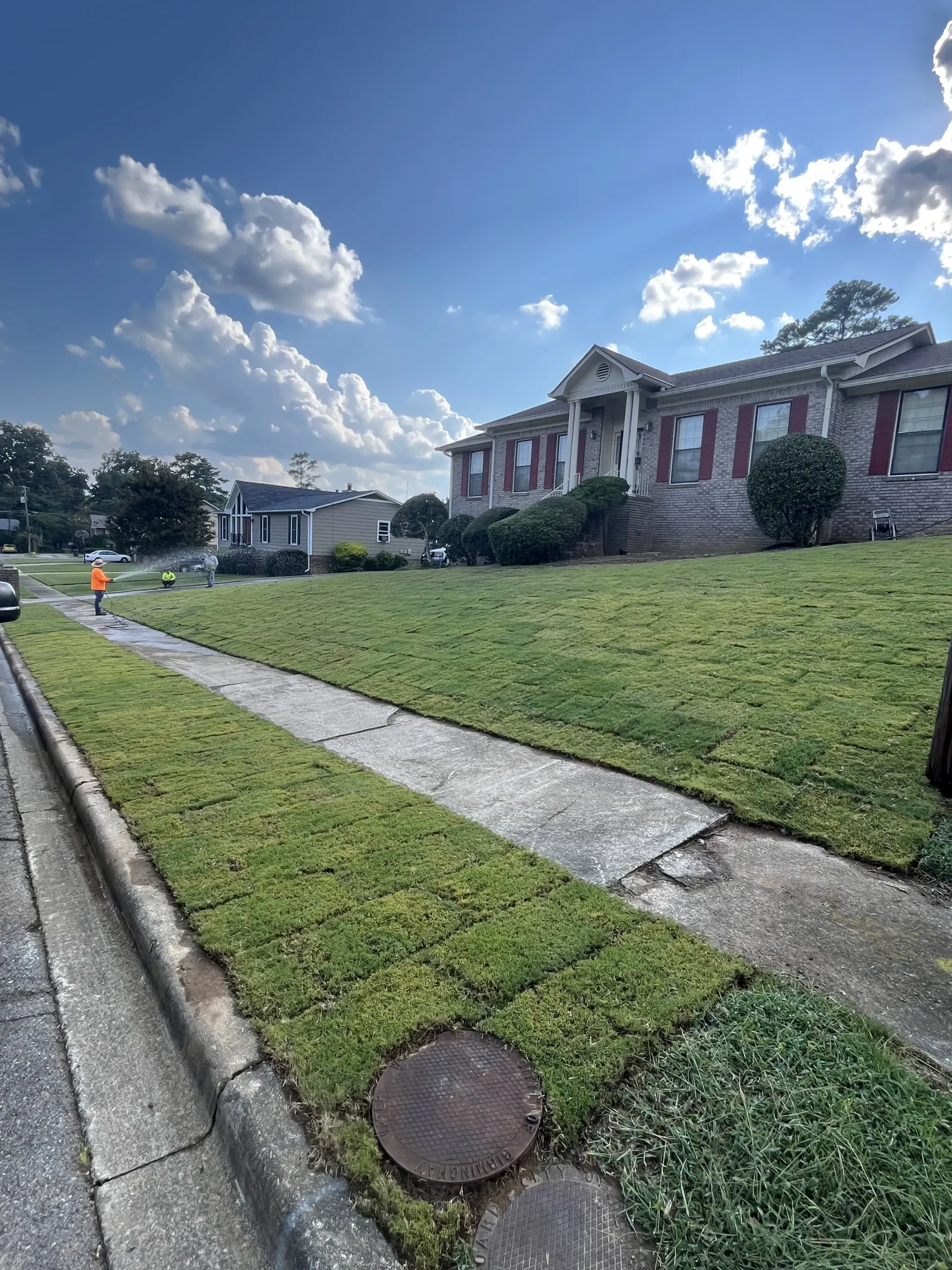 Sidewalk beside a grassy slope leading to a stone house under a bright blue sky with clouds.