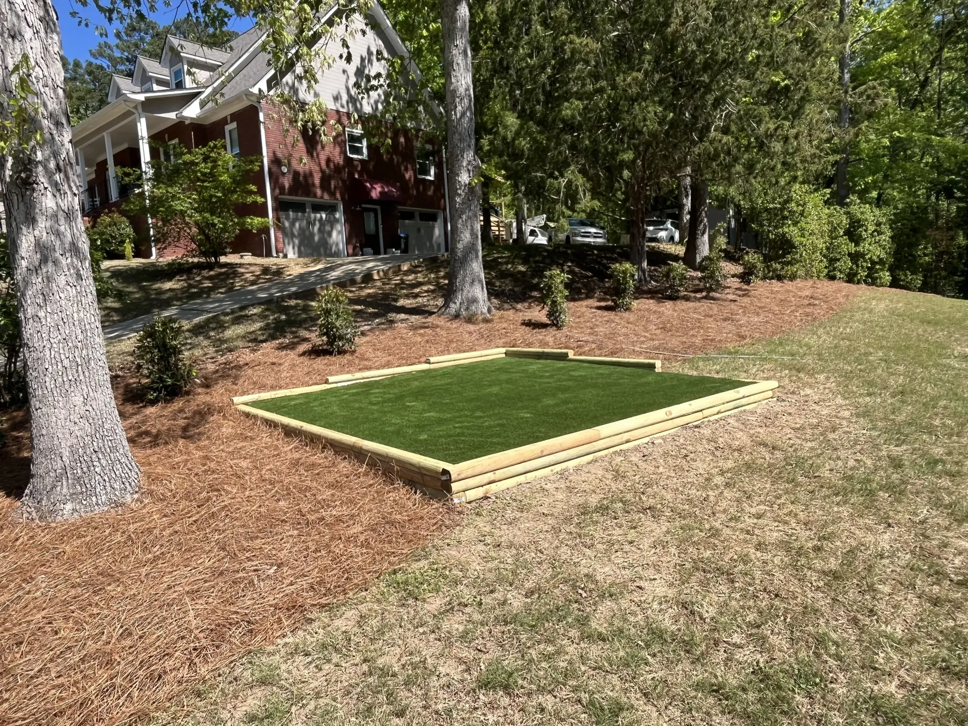 Raised square turf patch in a mulched yard beside trees and a house