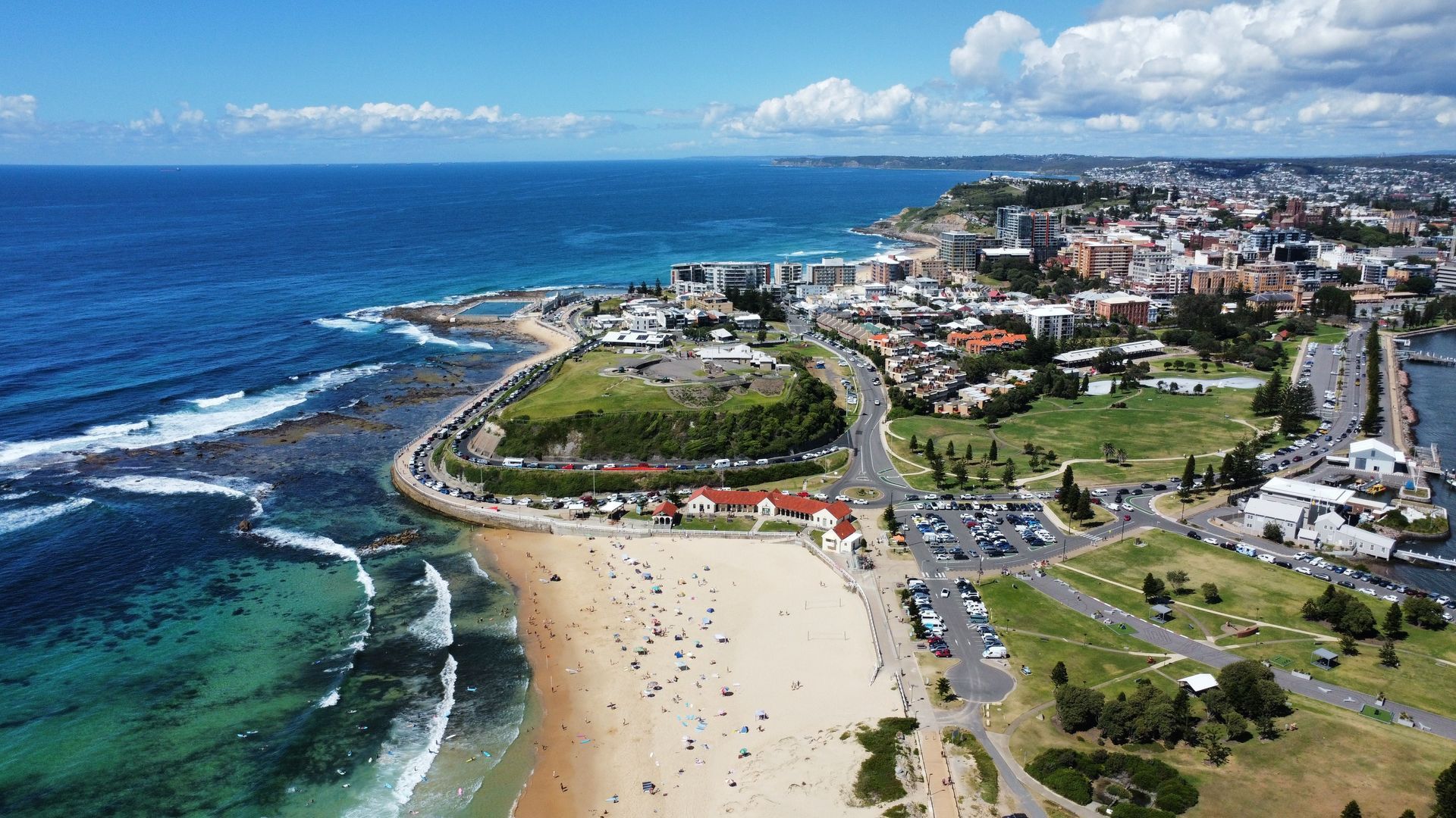 Aerial view of a coastal beach town with blue ocean, sandy shore, green park, and roads under a bright sky
