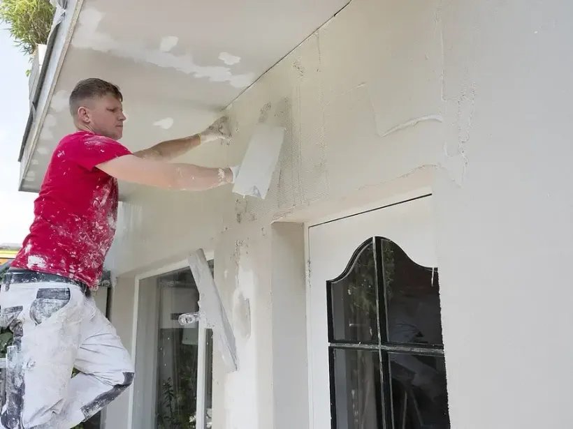 Person painting the exterior wall of a house with white paint while standing on a ladder