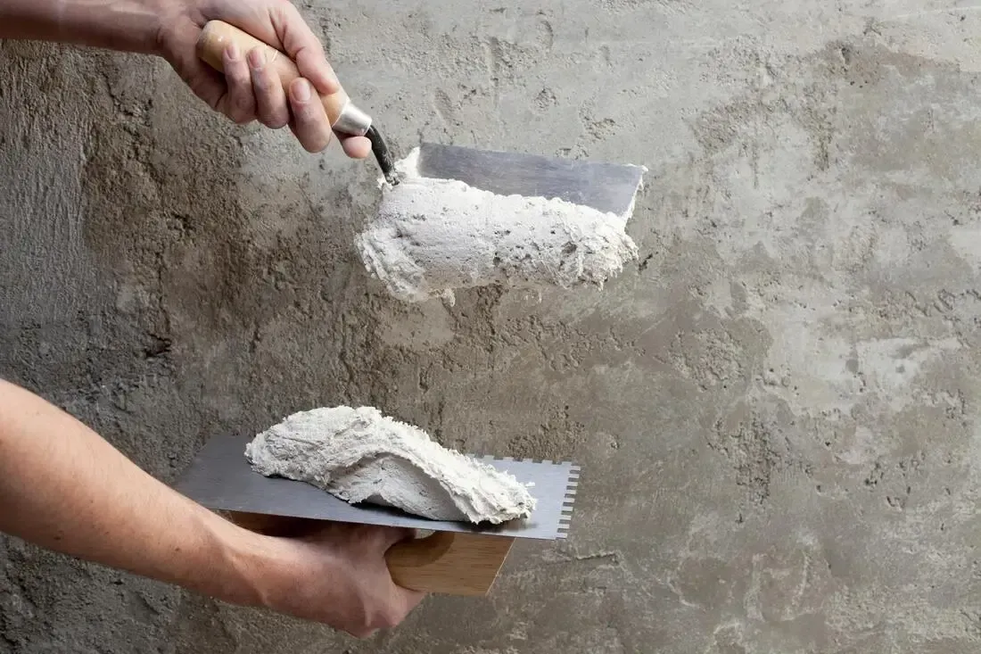 Hands using trowels to spread gray plaster on a concrete wall.