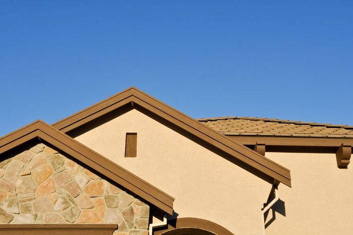 Tan stucco house roofline with stone accent wall against a clear blue sky