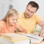 Adult and child reading together at a table, with open books and a pen.