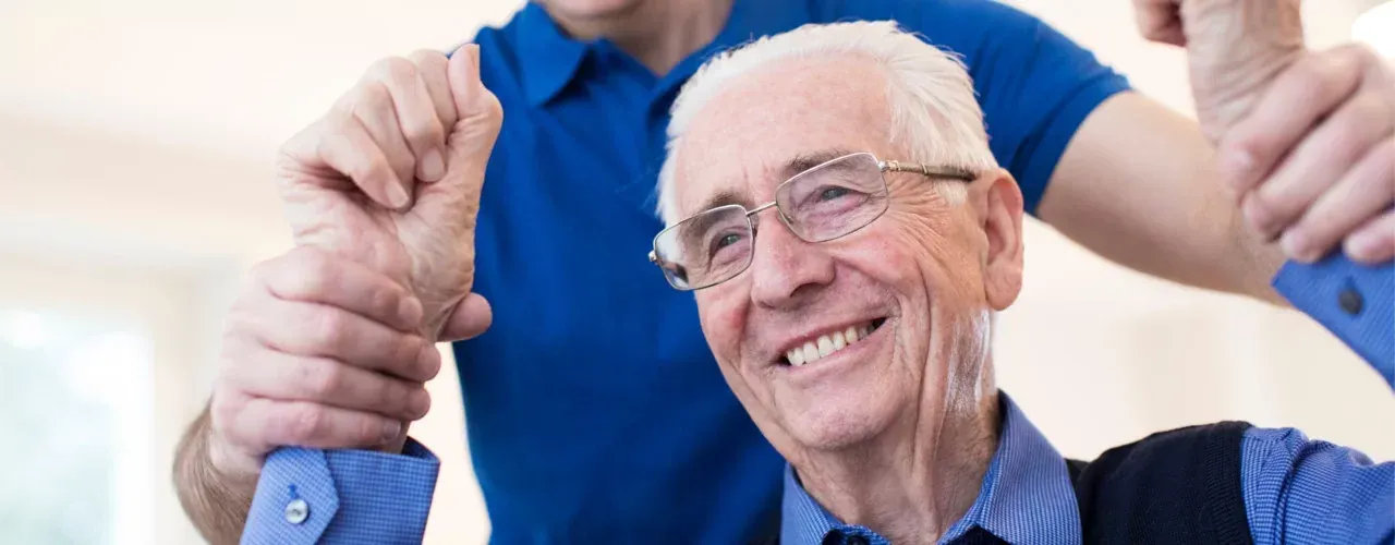 Senior man smiling while holding parallel bars during physical therapy with a trainer nearby