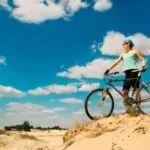 Cyclist standing on a sandy hill under a blue sky with clouds