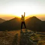 Silhouette of a person standing on a mountain peak at sunrise with one arm raised against golden sky