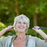 Smiling person with short white hair holding a garden tool handle outdoors in front of greenery