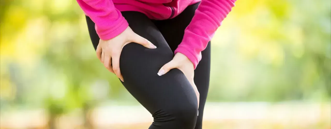 Person in a pink top holding their knee while standing outdoors, suggesting knee pain or injury